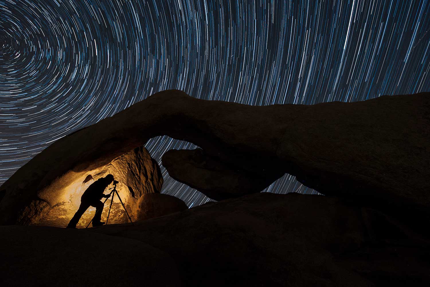 A photographers' silhouette against a rock formation photographing the stars with long exposure.