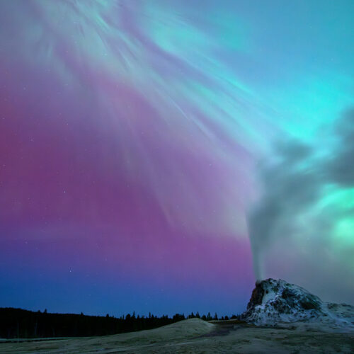 Yellowstone National Park, Wyoming A coronal aurora illuminates the landscape surrounding White Dome Geyser as it erupts under the night sky, casting a mystical rainbow glow over the scene.