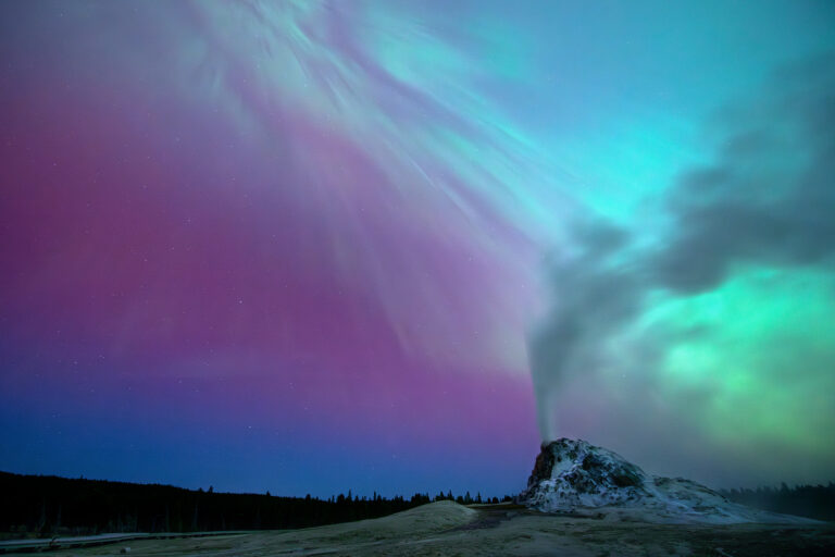 Yellowstone National Park, Wyoming A coronal aurora illuminates the landscape surrounding White Dome Geyser as it erupts under the night sky, casting a mystical rainbow glow over the scene.