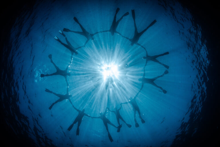 a circle of scuba swimmers centered around a light fixtured above, with photographer underneath the waters image looking like an eye
