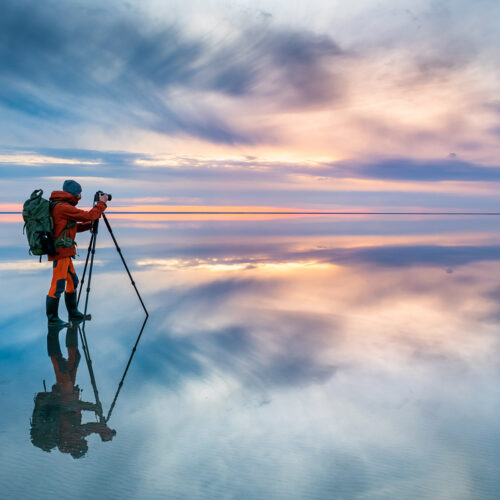 A nature photographer standing on an illuminated shallow lake capturing the sunset that is reflecting across the horizon.