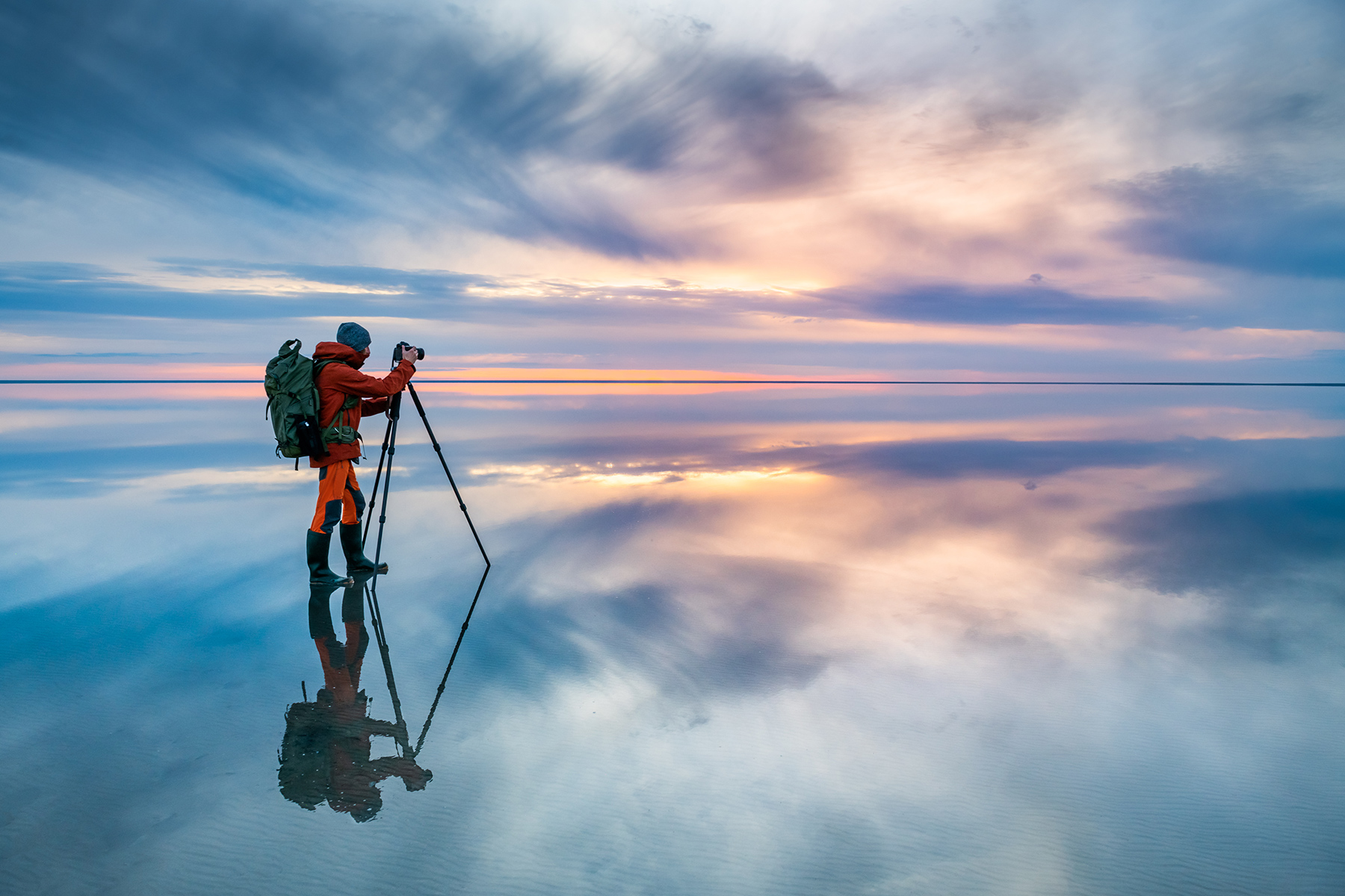 A nature photographer standing on an illuminated shallow lake capturing the sunset that is reflecting across the horizon.