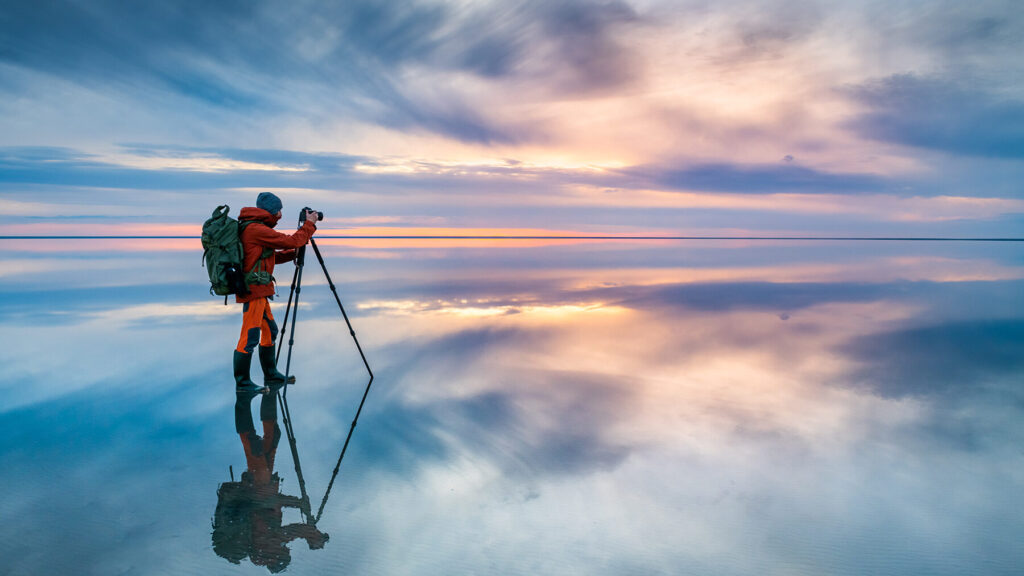 A nature photographer standing on an illuminated shallow lake capturing the sunset that is reflecting across the horizon.